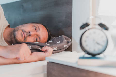 Sleepless man lying in bed and looking at alarm clock on bedside nightstand, selective focusの写真素材