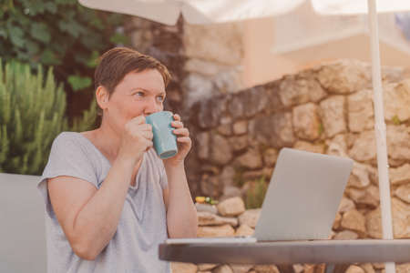 Woman drinking coffee while working from home terrace on laptop computer in summer morning