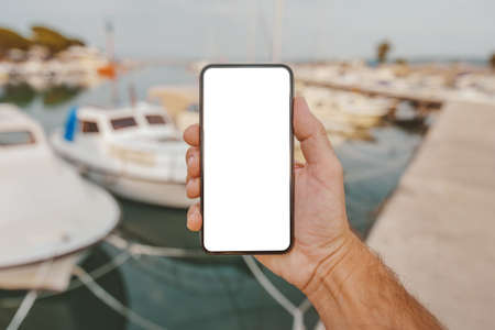 Fisherman holding smartphone with blank mock up screen in front of his boat in marina, selective focusの写真素材