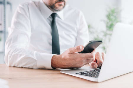 Entrepreneur using cellphone and laptop computer in bright business office interior, hand holding mobile smart phone, selective focusの写真素材
