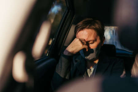 Businesswoman with severe headache pain symptoms and protective face mask at the backseat of a car, selective focusの写真素材