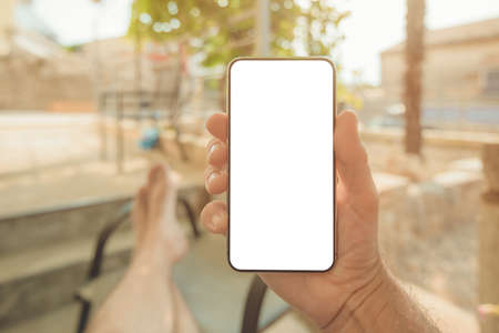 Relaxed man sitting on apartment terrace with feet on the table looking at mobile smart phone mockup screen, selective focusの写真素材
