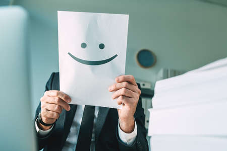 Happy businessman holding paper with smiling emoticon in office interior, selective focusの写真素材