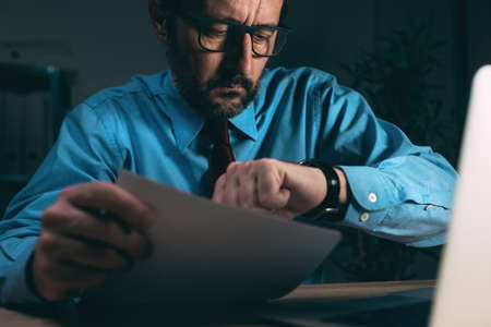 Working late night shift, businessman in dark office interior looking at his wristwatch, selective focusの写真素材