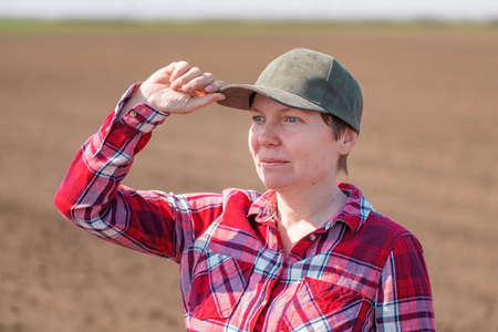 Portrait of female farmer with baseball cap and plaid shirt posing in plowed agricultural field prepared for seeding season, selective focusの写真素材