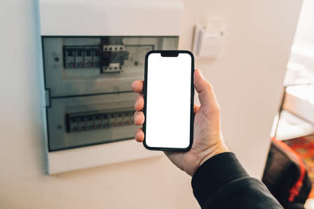 Electrician holding smartphone with blank mockup touch screen in front of circuit breaker box at...