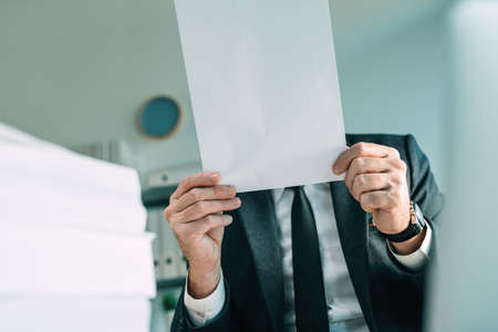 Unrecognizable business person holding blank white mockup paper in front of his face as copy space,...