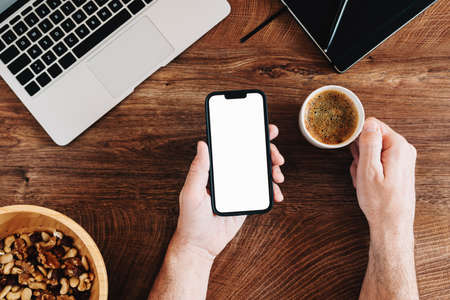 Freelancer using mobile phone with blank white mockup screen and drinking coffee at office desk, top...