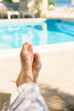 Barefoot male tourist relaxing by the outdoor swimming pool in seaside resort on sunny summer day, pov shot with selective focusの写真素材