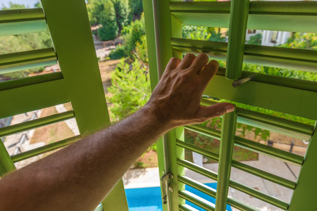 Male hand opening green wooden window shutters at seaside tourist resort in summer morning, selective focusの写真素材
