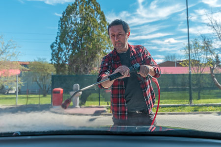 Man washing car with water gun in carwash self-service. Soap sud, wax and water drops covering vehicle window glass. Seen from the inside of the automobile.の写真素材