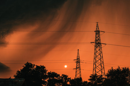 Dark ominous stormy clouds at orange summer sky, silhouettes of treetops and electricity pylon just...