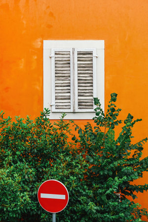 Colorful urban street scene - No Entry traffic sign next to an old orange building with closed white window and green treetop, selective focusの写真素材