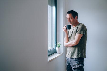 Pensive man drinking coffee in the morning by the apartment window while looking out and thinking. Caffeine dose for starting of the day. Selective focus.の写真素材