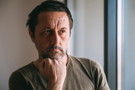 Serious caucasian mature adult male thinking by the apartment window, selective focus