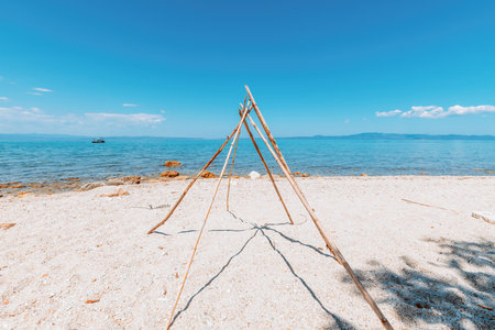 Rustic wooden tripod structure made of driftwood sticks on a sandy Greek beach with clear turquoise sea and blue sky. Serene coastal landscape.の写真素材