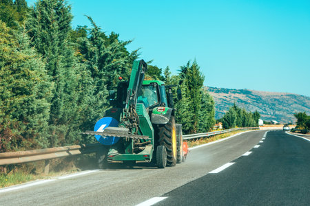 Tractor with hydraulic mower cutting roadside grass along a mountain highway on a sunny summer day. Selective focus.の写真素材