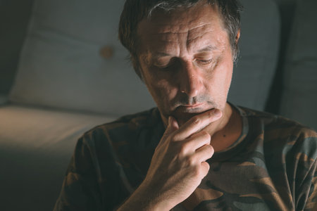 Pensive middle-aged man sitting indoors in low light, deep in thought with hand on chin, selective...