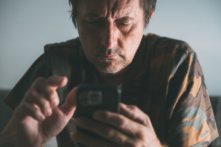 Focused middle-aged man using smartphone at home, intense concentration in low light, selective focusの写真素材