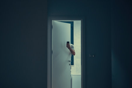 Man partially hidden behind door holding smartphone in dimly lit interior, mysterious atmosphere. Hand with smartphone visible through open door, moody low light interior scene with suspenseful feel.の写真素材
