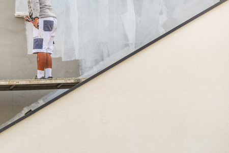 Male painter painting building facade with white color, renovation and maintenance work. Construction worker with roller applying fresh white paint on building exterior wall. Selective focus.の写真素材