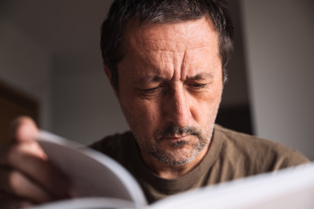 Thoughtful older man concentrating on reading a book indoors at home in living room. Selective focus.の写真素材