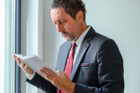 Mature businessman in formal suit with red necktie reading book by office window, concept of knowledge and learning. Selective focus.の写真素材
