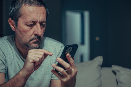 Tired mature man typing text message on smartphone in dark living room interior, using mobile phone with index finger, low key portrait in casual home setting. Selective focus.の写真素材