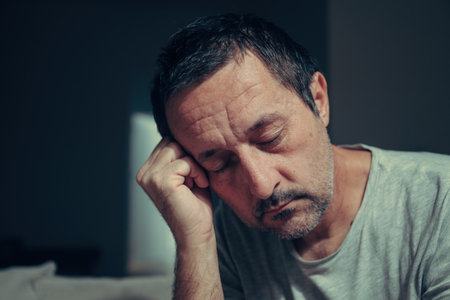 Sleepy tired man resting head on hand with eyes closed in dark low key living room interior at night. Selective focus.の写真素材