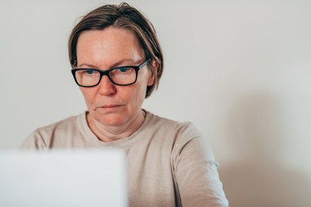 Serious female freelancer working on laptop at home office, mature brunette woman in casual attire and eyeglasses. Selective focus.の写真素材