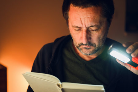Middle-aged man reading a book at night, holding a portable LED light for illumination, quiet focus...
