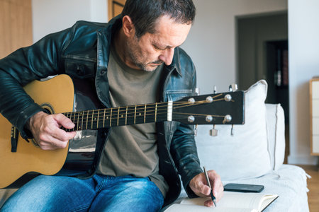 Mature man at home playing acoustic guitar and writing lyrics in notebook, concept of creativity,...