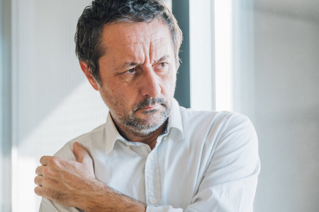 Unshaved mature businessman suffering from severe shoulder pain, holding arm in discomfort. Selective focus.の写真素材
