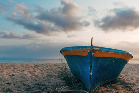 Old blue fishing boat with orange trim resting on sandy beach at sunset. Cloudy sky over calm sea in the background. Peaceful, rustic coastal scene. Selective focus.の写真素材