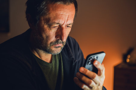 Tired unshaved mature man using smartphone in dark room illuminated by warm low key light. Selective focus.の写真素材
