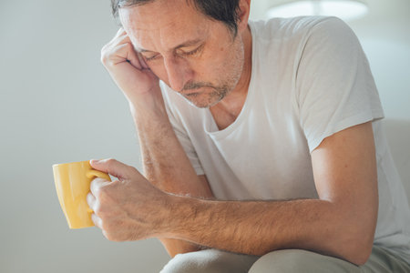 Reflective adult male with tired look holding yellow mug, sitting in cozy chair at home in morning light. Selective focus.の写真素材