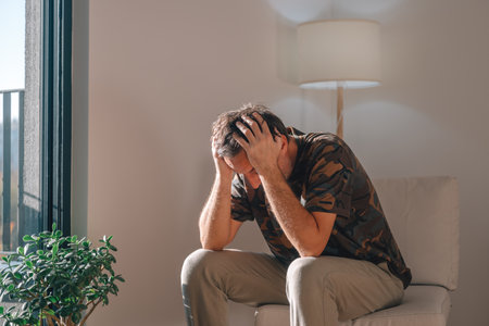 Depressed man leaning forward with hands on his head, struggling with anxiety or bad news in a bright, quiet living room. Selective focus.の写真素材
