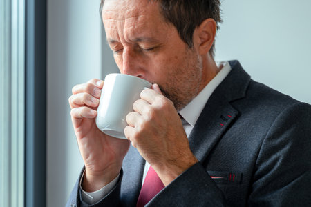 Calm businessman enjoying first sip of morning coffee from white cup, serene office interior. Selective focus.の写真素材