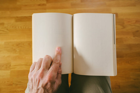 POV of person holding open blank book mockup with finger pointing at empty page over wooden floor...