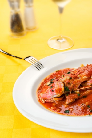 Red Mullet seafood italian traditional gourmet recipe with tomato, garlic, olive oil with wine glass, salt and pepper on a yellow tableclothの写真素材