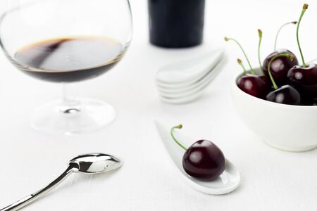 Single cherry on a white china spoon, a cherries group in a small round white bowl and a sherry glass and bottle on backgroundの写真素材