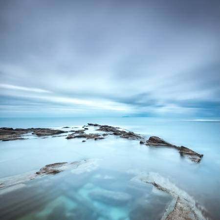 Dark rocks in a blue ocean under cloudy sky in a bad weather. Long exposure photographyの写真素材