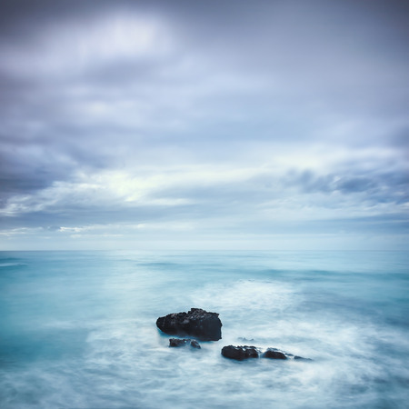 Dark rocks in a blue ocean under cloudy sky in a bad weather. Long exposure photographyの写真素材