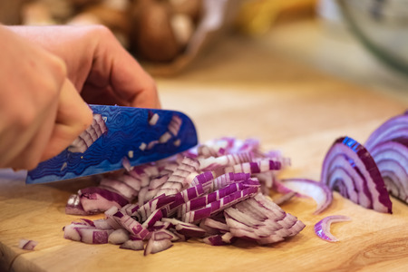 Hands chopping red onion with a blue knife on wood cutting board.の写真素材