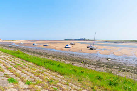 From the top of a sloping grass bank out across the estuary at low tide, where small yachts and motor boats sit on the sand, out across the salt marshes to the horizonの写真素材