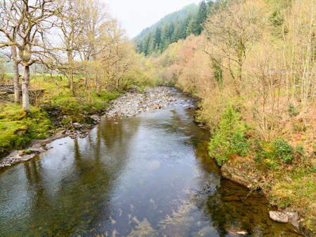 The shallow, clear, River Mawddach in Gwynedd, Wales, flows gently over rocks and between banks lined with trees and fields.の写真素材