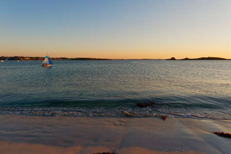 As the sun sets on a beach at Landeda, in Brittany, a small sailing boat is out on the calm waterの写真素材