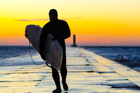 GRAND HAVEN , MI - November 24, 2017: A surfer wearing a full-body wetsuit walks with his surf board on the north pier as the sun sets over Lake Michigan.のeditorial素材