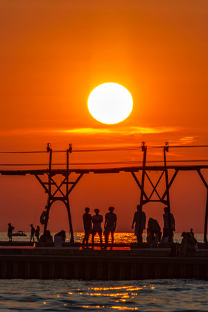 People silhouetted against a dark orange sky as the sun sets behind the Grand Haven pier in Michiganの写真素材