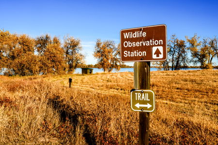 A sign points to a wildlife observation station, the wood hut in the background, at Barr Lake State Park northeast of Denver, Colorado, on a fall morning. A bird watcher contemplates the scene.の写真素材